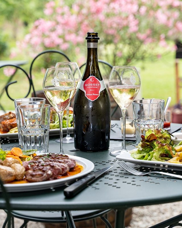 An outdoor dining setting with a bottle of Gosset champagne on a metal table surrounded by full glasses of wine, water glasses, and plates of food