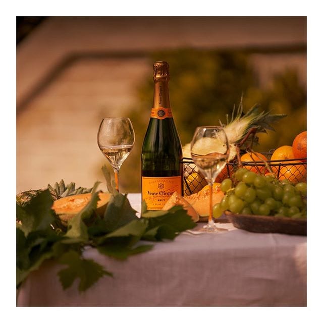 A bottle of Veuve Clicquot champagne, two wine glasses partially filled with champagne, and a rich assortment of fruits