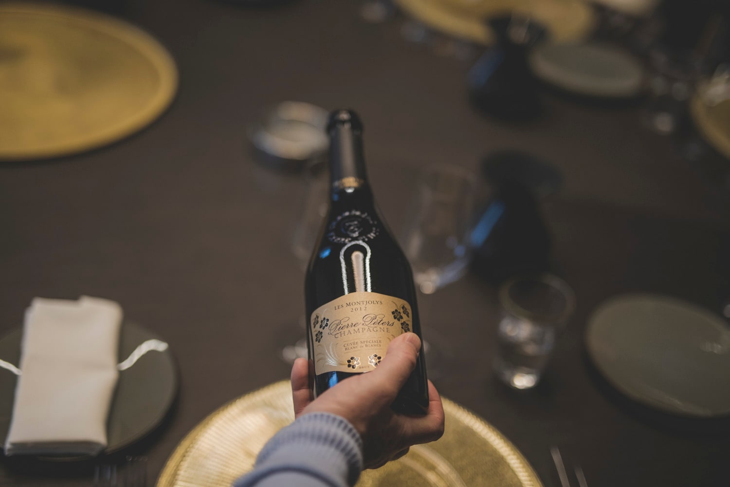 A bottle of Pierre Peters champagne, with a blurred dining table setting in the background