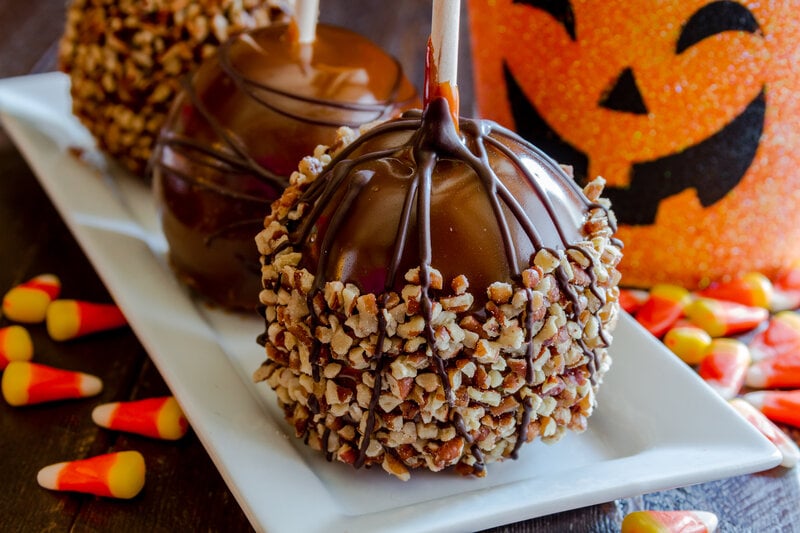 Homemade caramel apples with nuts and chocolate drizzle sitting on white plate with halloween pumpkin bucket and candy corn